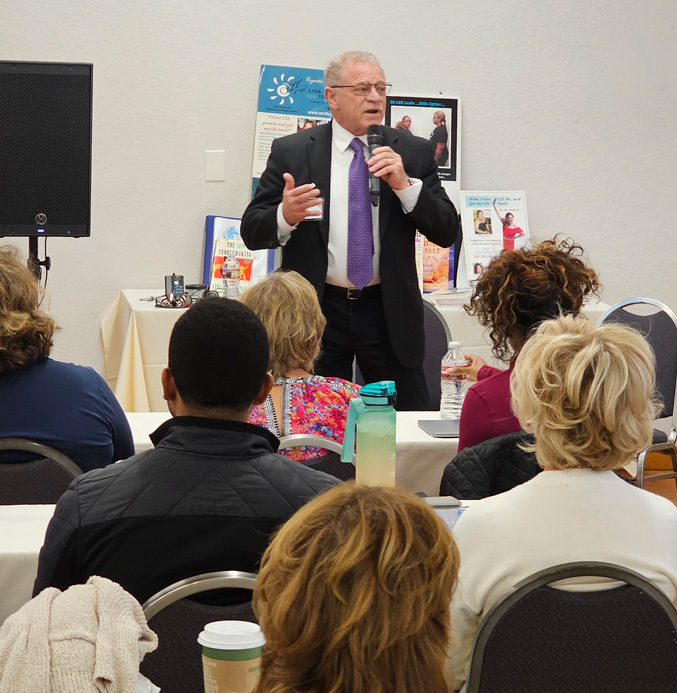 A man in a suit speaks into a microphone in front of an audience seated at tables in a conference room, with posters and informational materials about hypnosis training visible in the background.