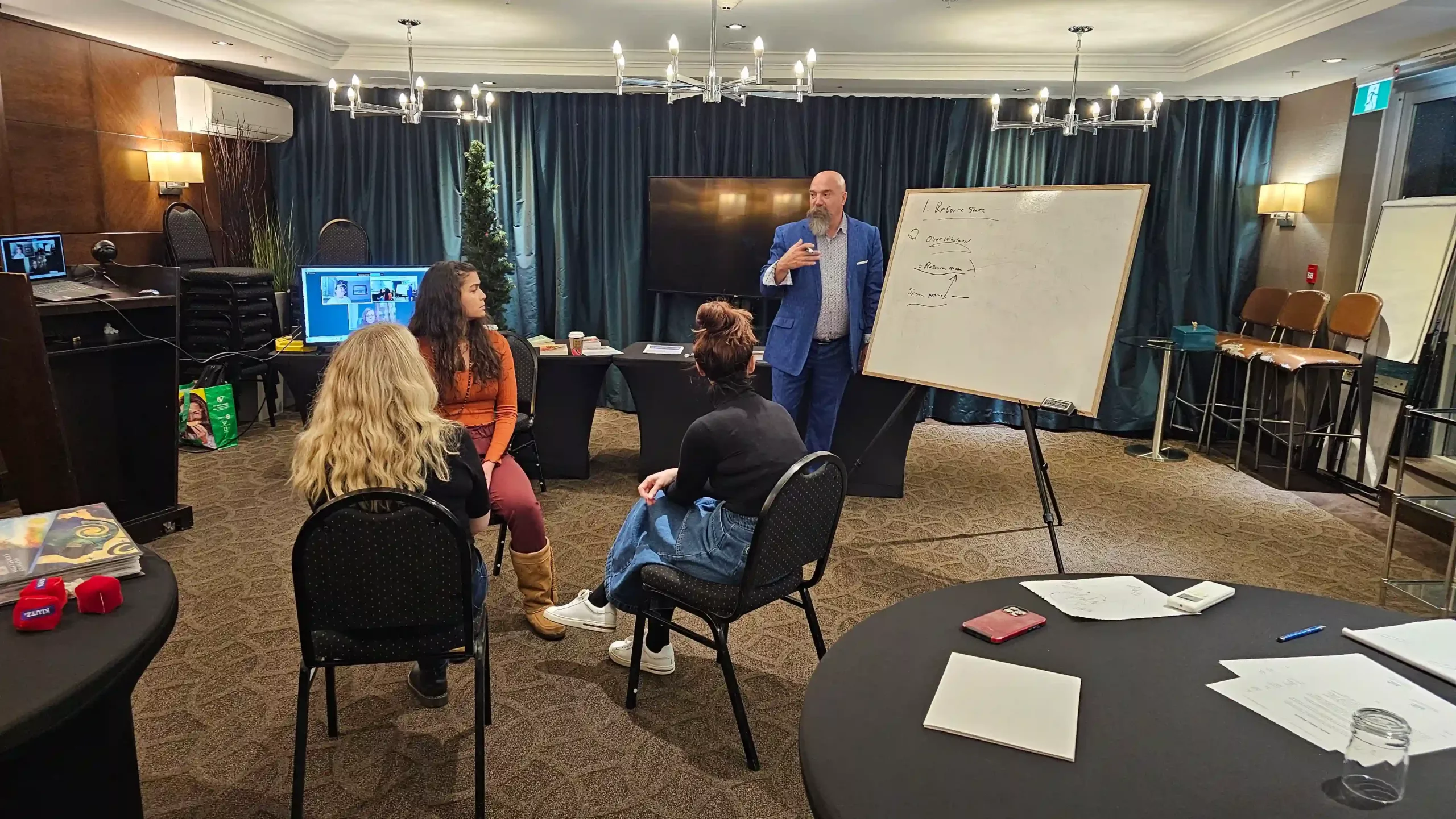 A man in a blue suit speaks to three women seated in a conference room, with a whiteboard, tables, and hypnosis training materials visible.