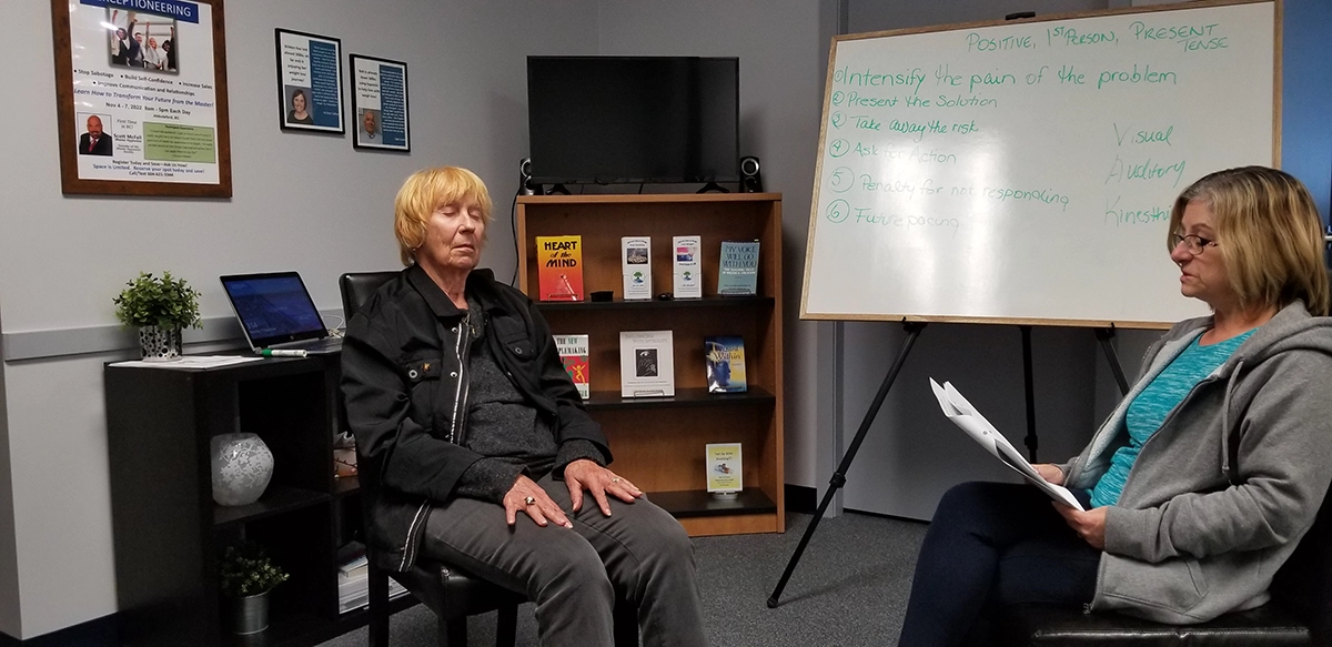 Two women sit facing each other in an office during hypnosis training; one has her eyes closed while the other holds papers. A whiteboard with notes and a bookshelf are visible in the background.