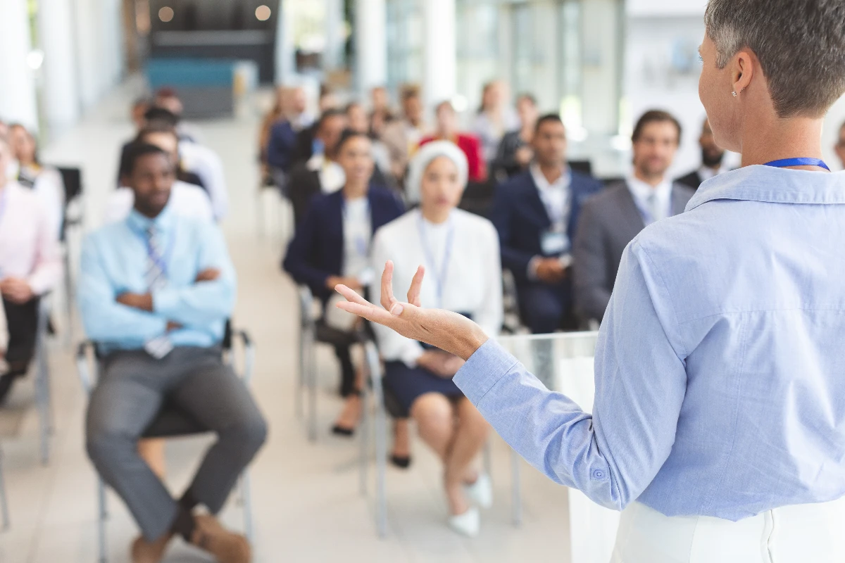A person stands at a podium speaking to an audience seated in rows, in a bright, modern conference room.