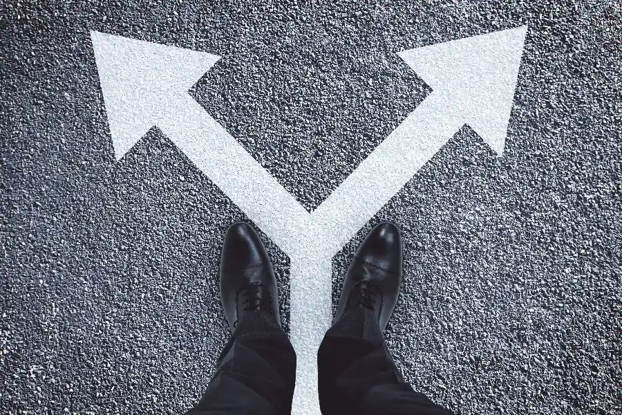A person in black dress shoes stands at a fork in the road with two large white arrows pointing left and right on the asphalt.