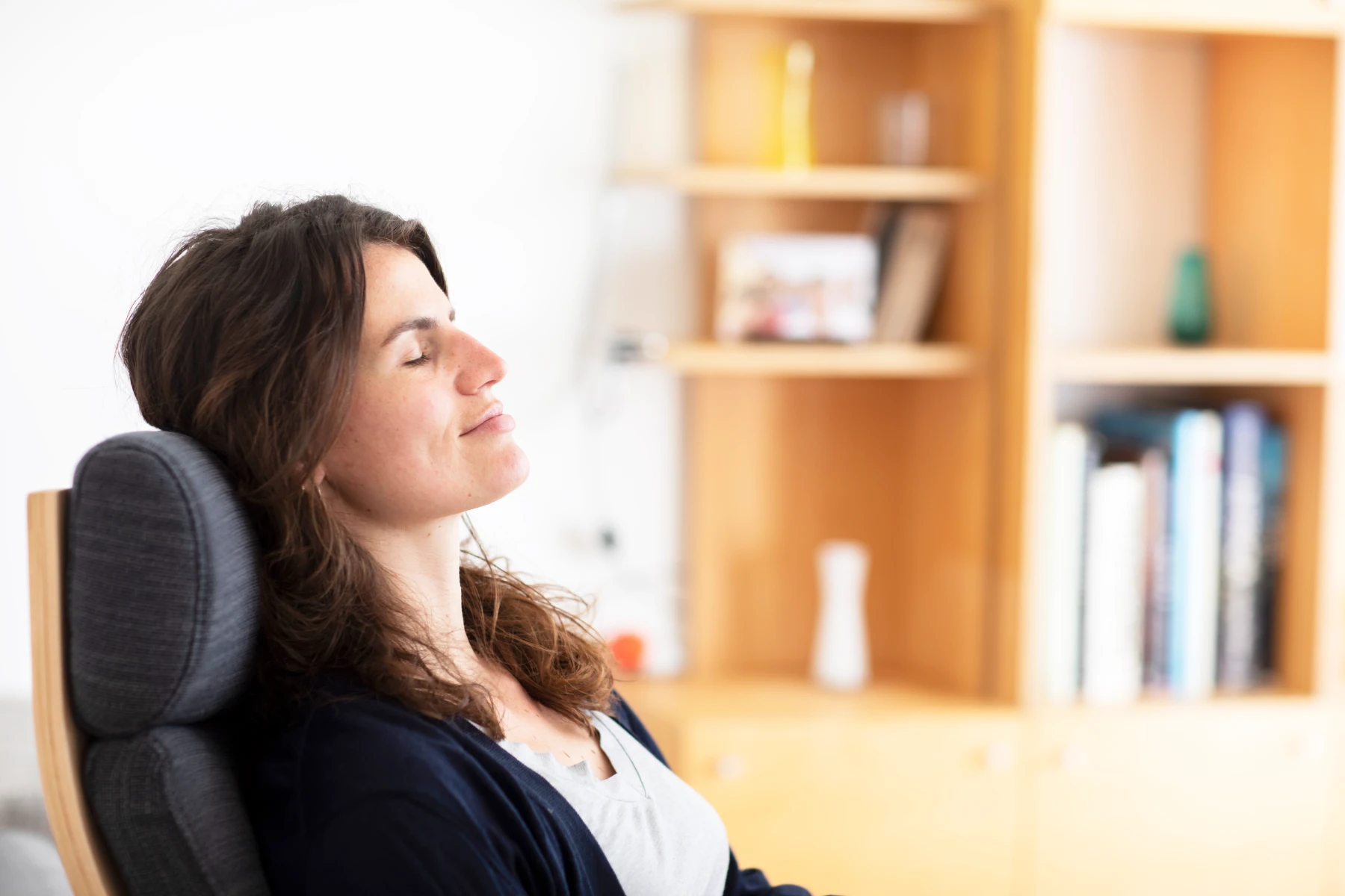A woman sits in a chair, eyes closed and smiling slightly, appearing relaxed. A wooden bookshelf with books and decor is in the background.