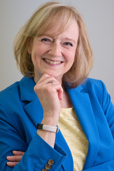 angela-nyhuis A woman with blonde hair wearing a blue blazer and yellow top smiles at the camera, resting her chin on her hand.
